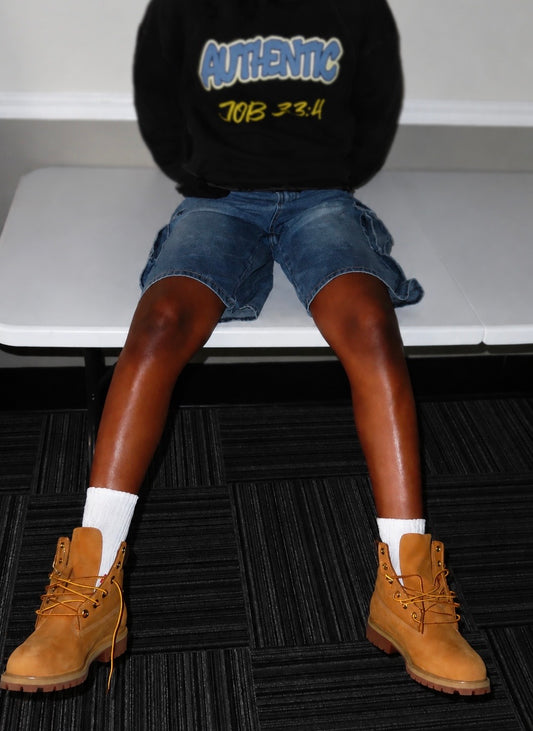A person seated on a white bench wearing a black sweatshirt with blue and yellow 'AUTHENTIC JOB 324' text, blue denim shorts, white socks, and tan lace-up boots on a dark patterned floor.