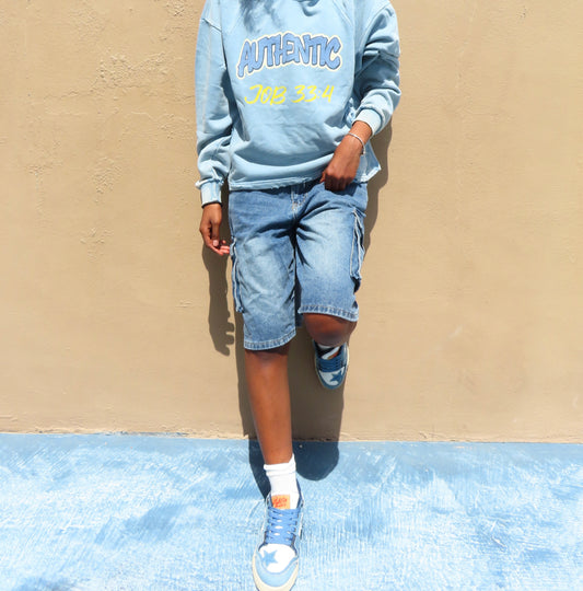 A person wearing a light blue sweatshirt with 'AUTHENTIC' and '2008-2014' printed on the chest, paired with denim shorts and blue sneakers, standing against a beige wall.
