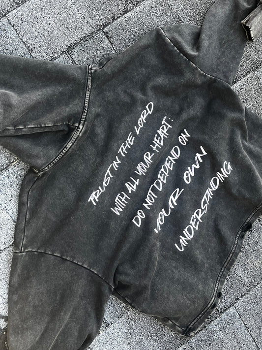 A dark gray acid-wash cropped hoodie with white printed text reading 'TRUST IN THE LORD WITH ALL YOUR HEART: DO NOT DEPEND ON YOUR OWN UNDERSTANDING', laid flat on gray stone pavers.