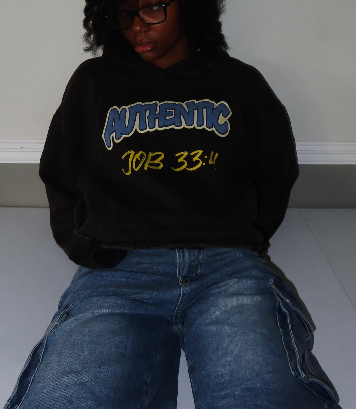 A person wearing a black hoodie with 'AUTHENTIC' in blue and yellow text, paired with blue jeans, sitting against a plain wall.
