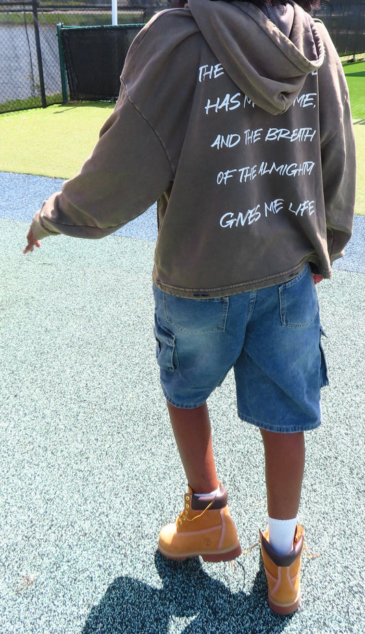 A person viewed from behind wearing a brownish-gray oversized hoodie with white handwritten text on the back, blue denim shorts, white socks, and tan work boots on a blue-gray outdoor playground surface.
